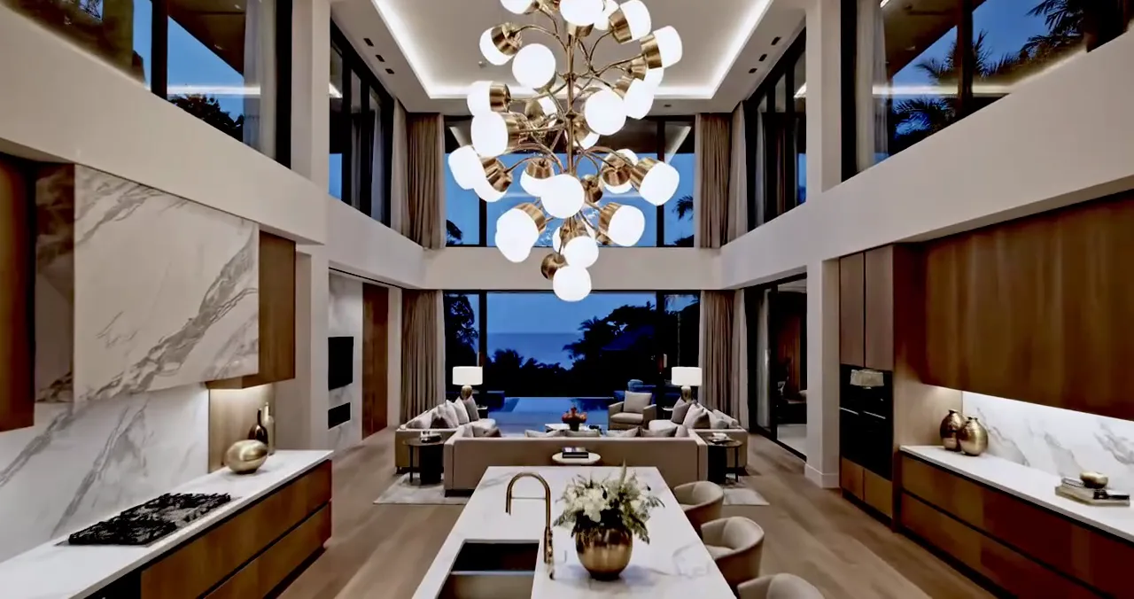 Double-height villa living room with a marble wall, a brass chandelier and a kitchen island, open to the pool